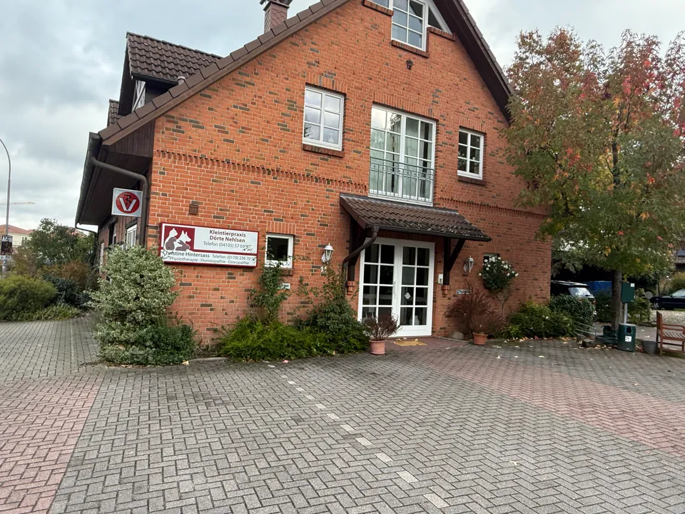 Giebelseite eines Backsteinhauses mit weißer Eingangstür und Schild der Kleintierpraxis, daneben ein Baum mit Herbstlaub