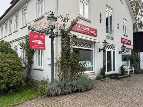Weißes Geschäftshaus mit roten Haferkamp-Immobilien-Schildern, Laterne mit Hängeschild, Schaufenster und Lavendelbeet auf gepflastertem Vorplatz