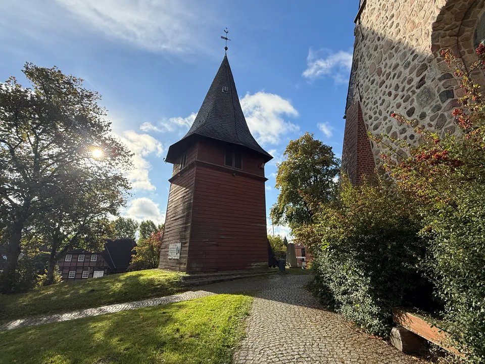 Roter Holzglockenturm mit dunkler Schieferspitze auf einem Kirchhof, links ein Fachwerkhaus im Gegenlicht