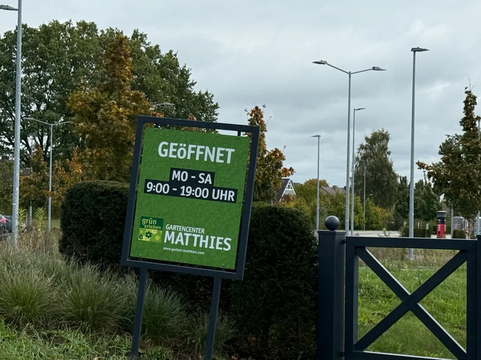 Grünes Schild mit Öffnungszeiten des Gartencenters Matthies, Montag bis Samstag 9 bis 19 Uhr, neben Hecke
