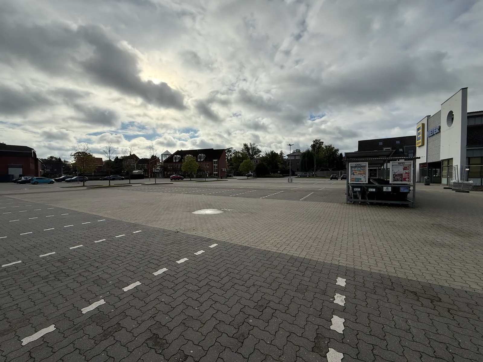 Weitläufiger gepflasterter Parkplatz mit Einkaufswagenunterstand, rechts Supermarkt mit EDEKA-Logo, dahinter Backsteinhäuser und Bäume