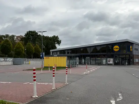 Parkplatz mit rot-weißen Pollern vor einer Lidl-Filiale mit Glasfassade und gelber DHL-Packstation unter bewölktem Himmel
