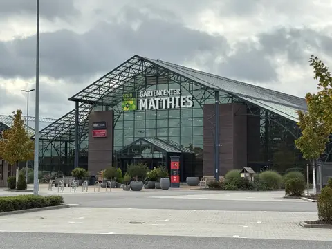 Glasfassade des Gartencenters Matthies mit Eingang, Pflanzkübeln, Fahrradbügeln und leerem Parkplatz unter bewölktem Himmel