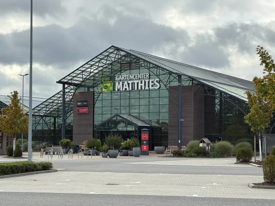 Glasfassade des Gartencenters Matthies mit Eingang, Pflanzkübeln, Fahrradbügeln und leerem Parkplatz unter bewölktem Himmel