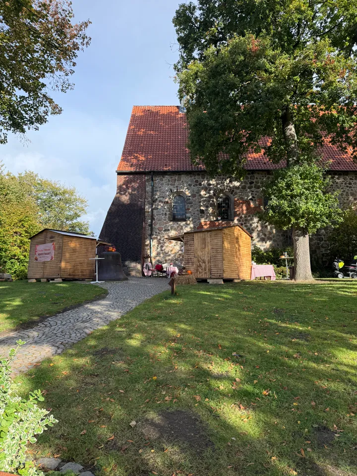 Holzbuden mit Herbstmarkt-Banner und eine alte Glocke vor einer Feldsteinkirche mit rotem Ziegeldach