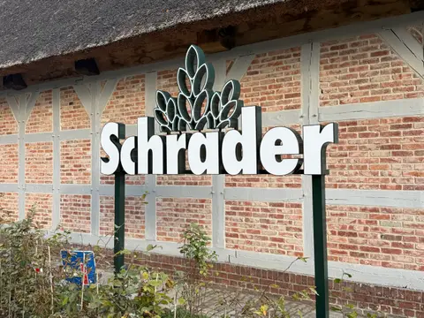 Weißer Schrader-Schriftzug mit Ährenlogo auf Pfosten vor einer Fachwerkwand mit Reetdach