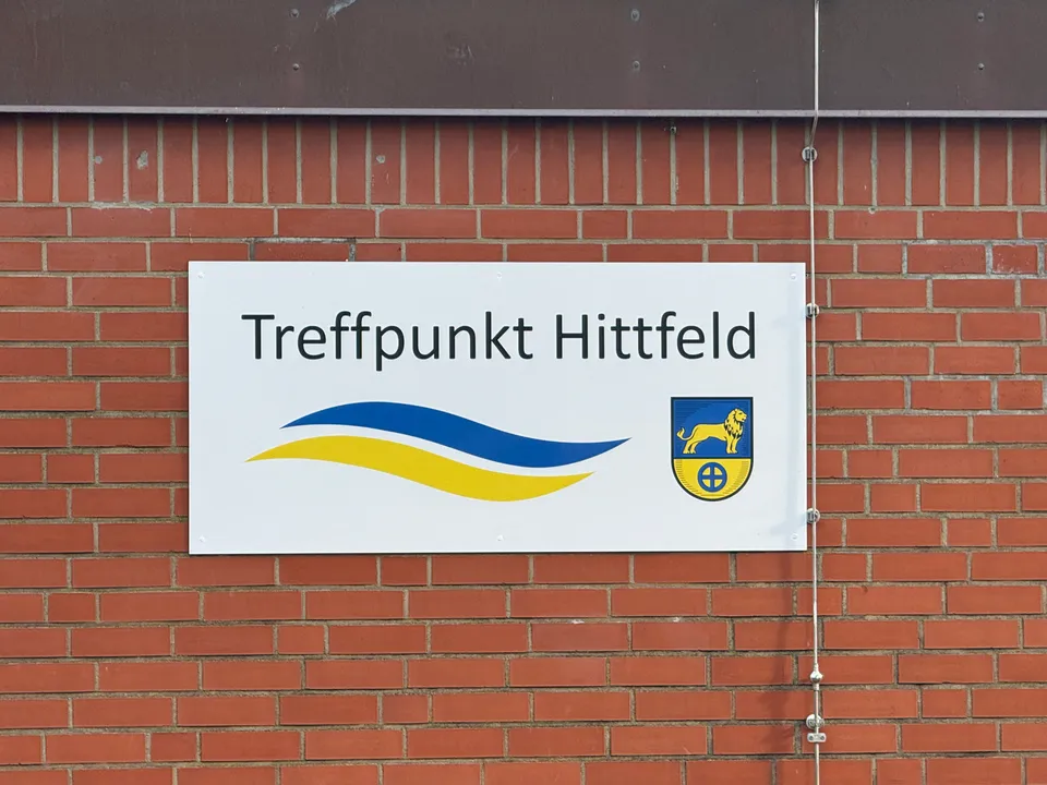 Weißes Schild „Treffpunkt Hittfeld“ mit blau-gelbem Wellenlogo und Löwenwappen an roter Klinkerwand